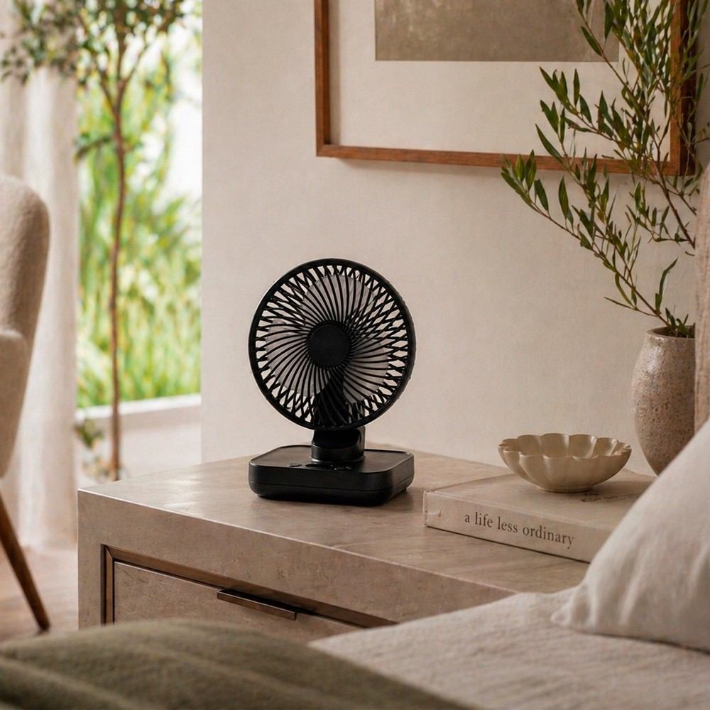 Un ventilador portátil negro está sobre una mesita de noche junto a un libro y un cuenco.