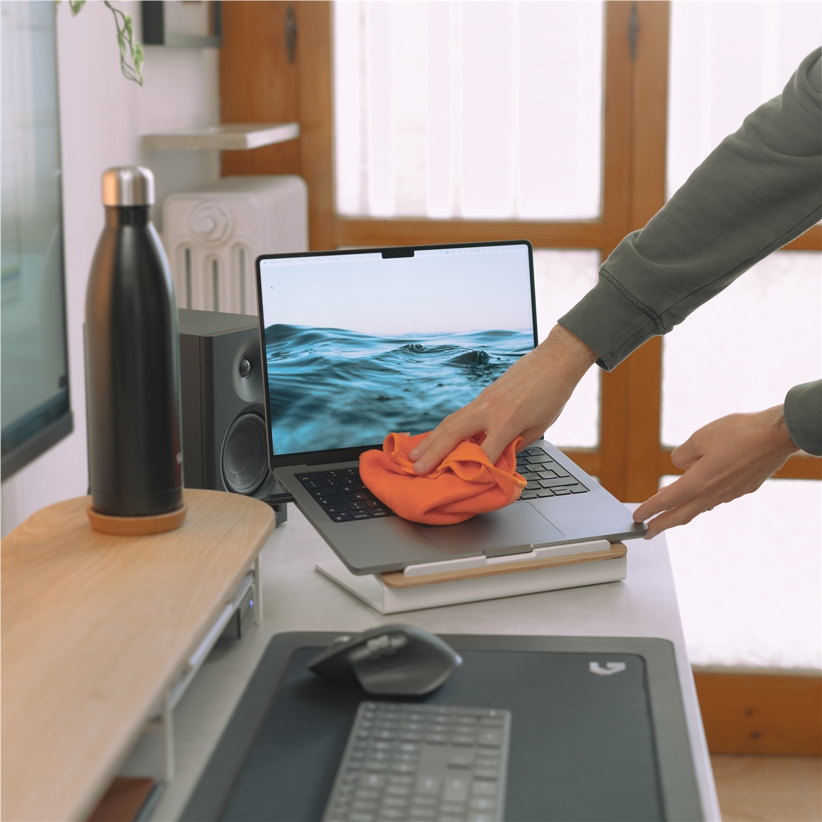 Persoon maakt laptop schoon met oranje doek, luidspreker en waterfles nabij.