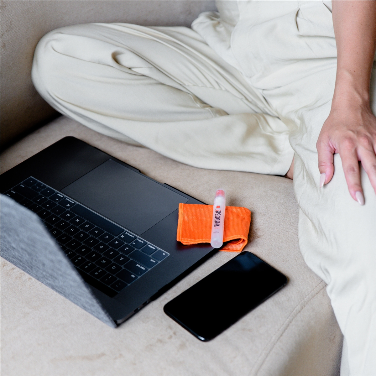 Person sitzt auf Sofa mit Laptop, Handy, Wischtuch und Reinigungsmittel.