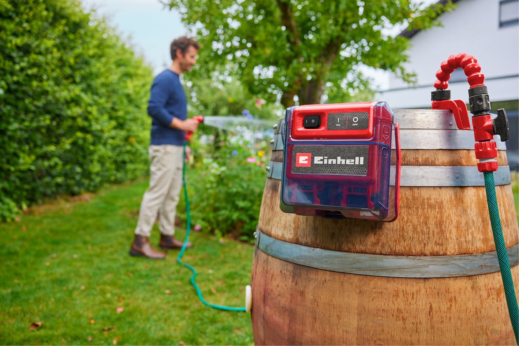 Mann wässert Garten mit Schlauch. Rote Pumpe auf Fass. Grüner Schlauch.
