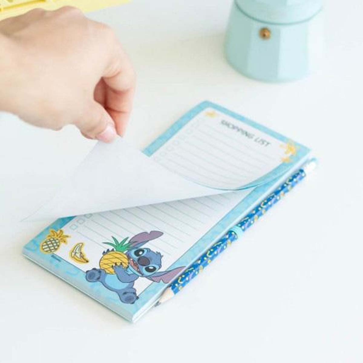 Ein Notizblock mit Stitch, der eine Ananas hält. Eine Hand blättert eine Seite um.