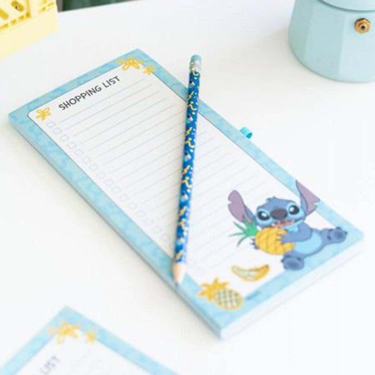 Ein Notizblock mit Einkaufsmotiv und Stift. Stitch hält eine Ananas.