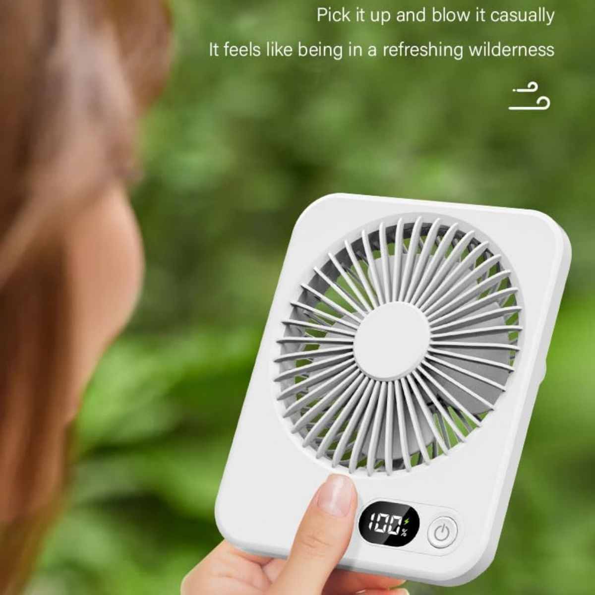 Eine weiße Hand hält einen kleinen weißen Ventilator vor unscharfem grünen Hintergrund.