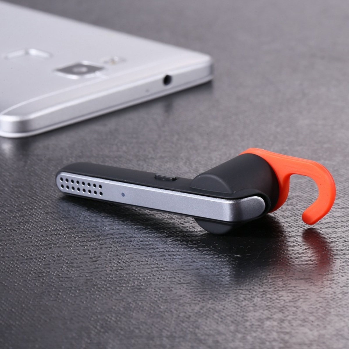 Ein schwarzes und oranges Headset liegt neben einem silbernen Smartphone auf einem dunklen Tisch.