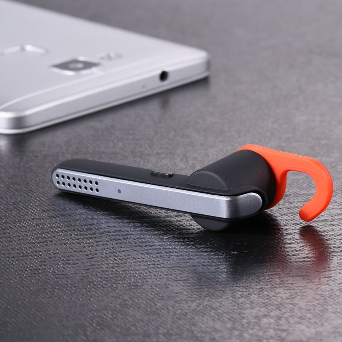 Ein schwarzes und silbernes Headset mit orangefarbenem Haken liegt neben einem Handy.