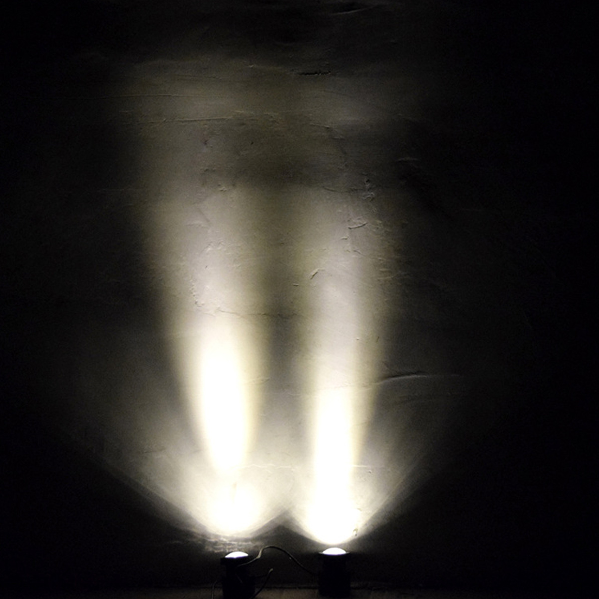Zwei Bodenscheinwerfer beleuchten eine dunkle Wand mit hellen Lichtstrahlen.