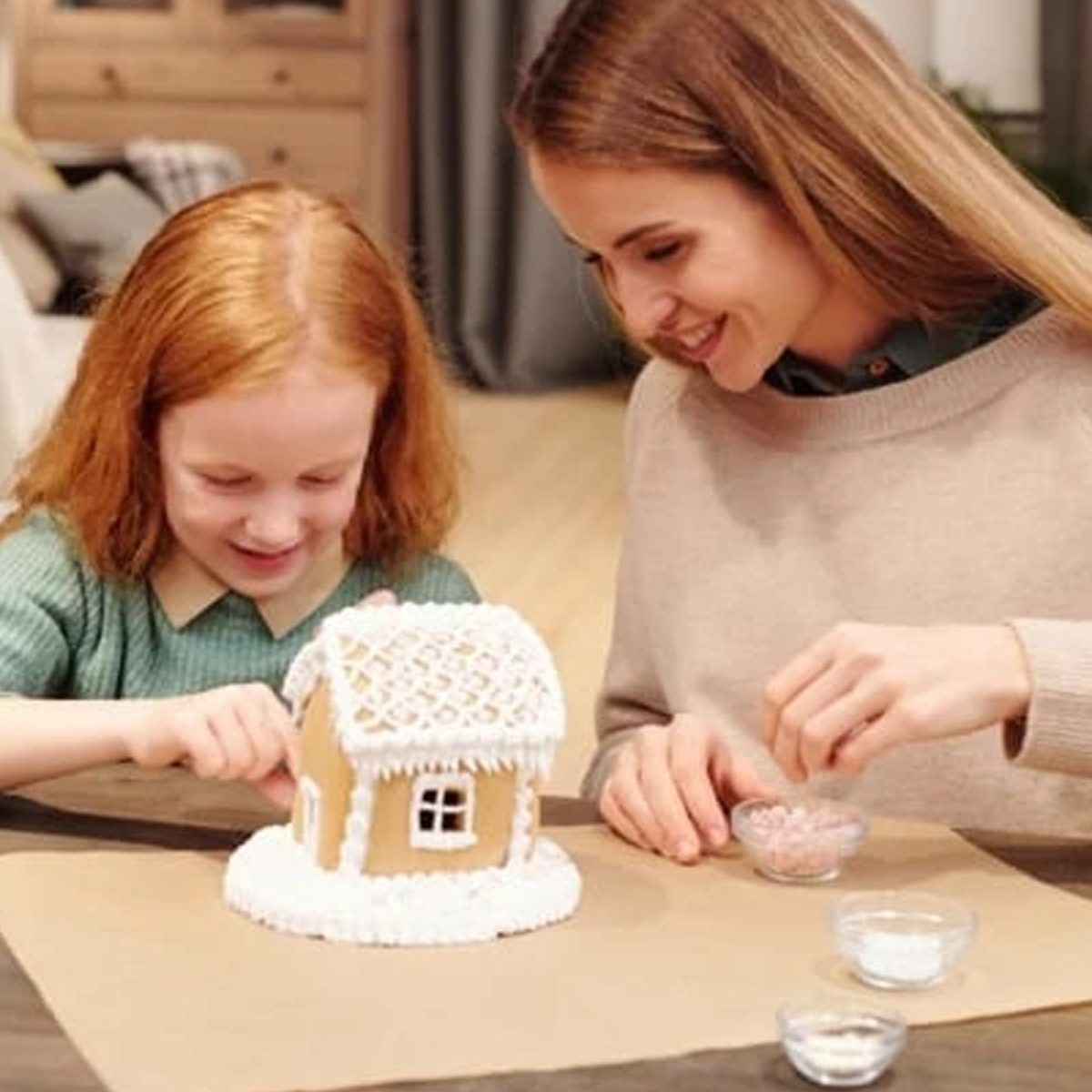 Ein Mädchen und eine Frau verzieren ein Lebkuchenhaus mit weißer Glasur und rosa Streuseln.