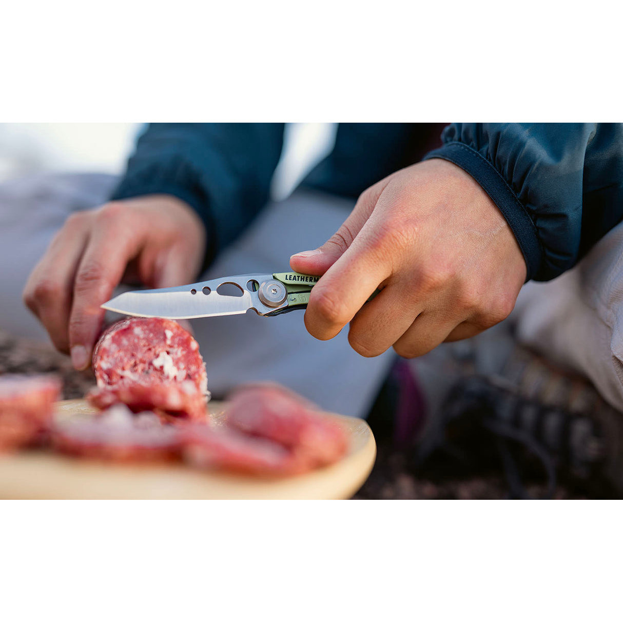 Person schneidet Salami mit einem Leatherman Multitool auf einem Holzbrett im Freien.