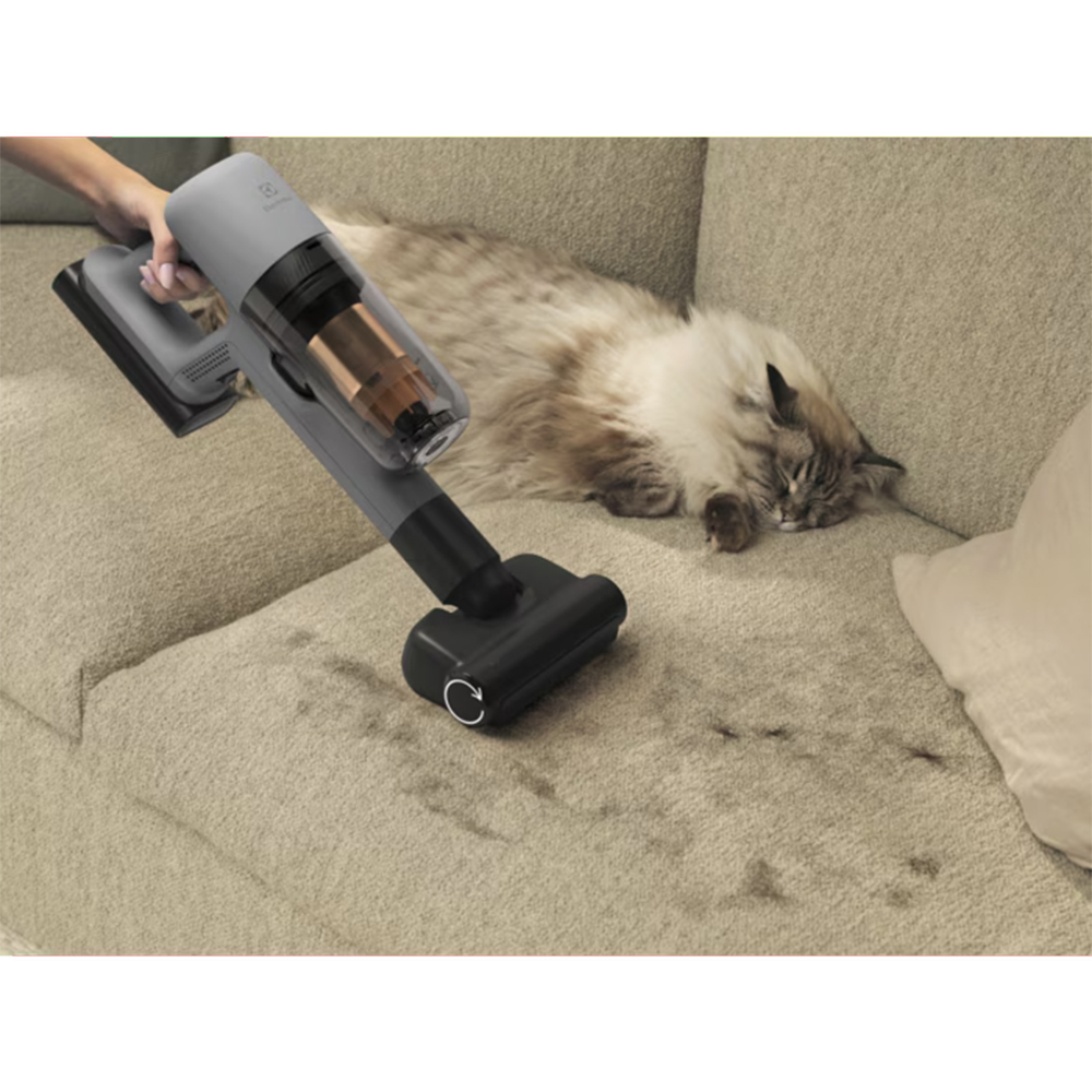 Una mano usa un aspirapolvere grigio su un divano dove dorme un gatto.