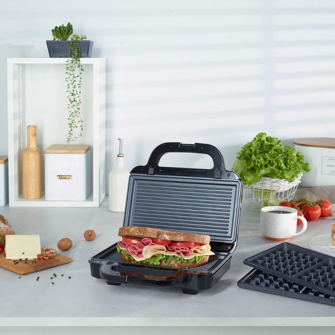 Açık tost makinesinde bir sandviç var. Tezgahta malzemeler ve mutfak gereçleri.