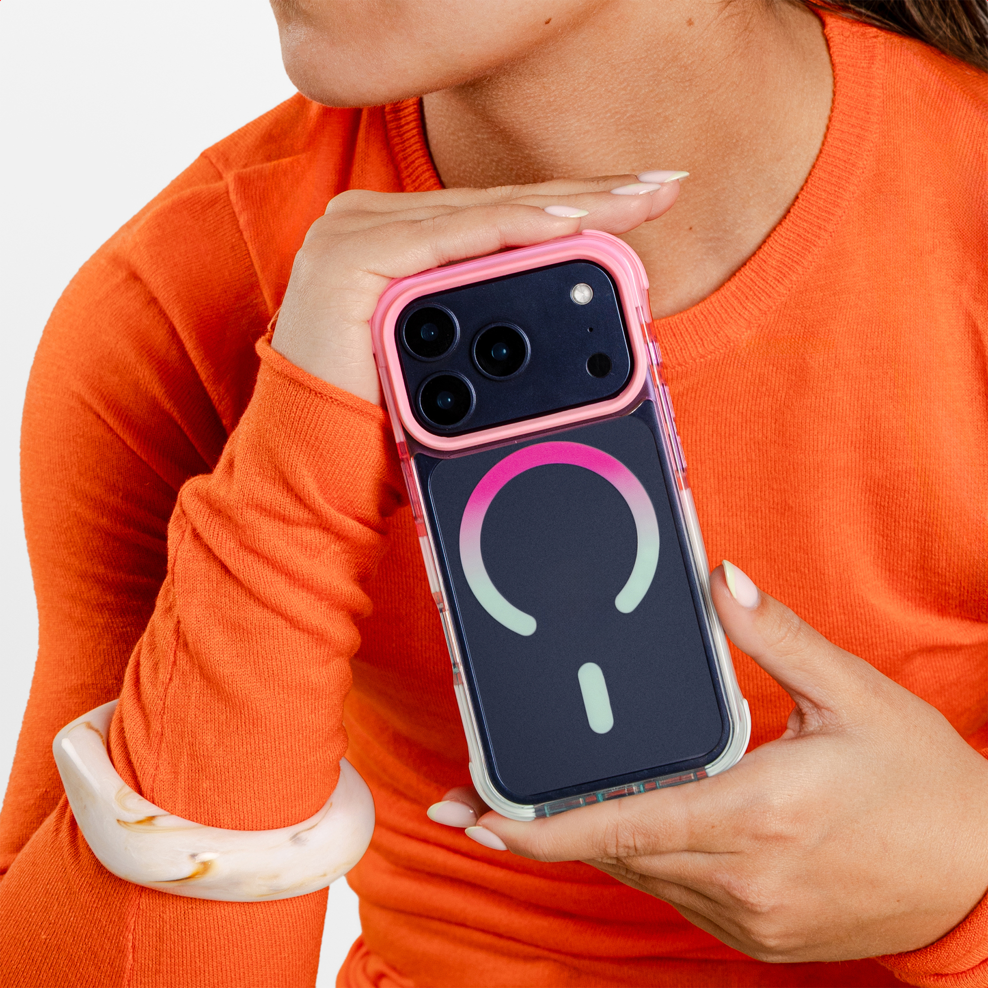 Een persoon in een oranje trui houdt een donkere telefoon met roze rand vast. Een armband is zichtbaar.