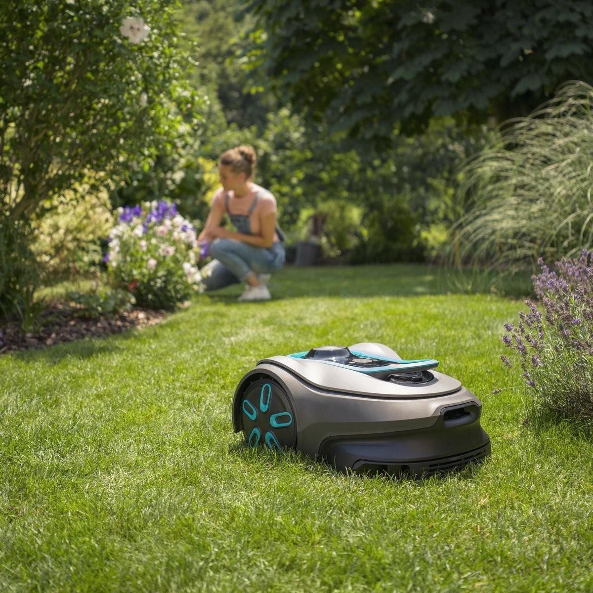 Un robot cortacésped está en un césped verde mientras una mujer se arrodilla al fondo.