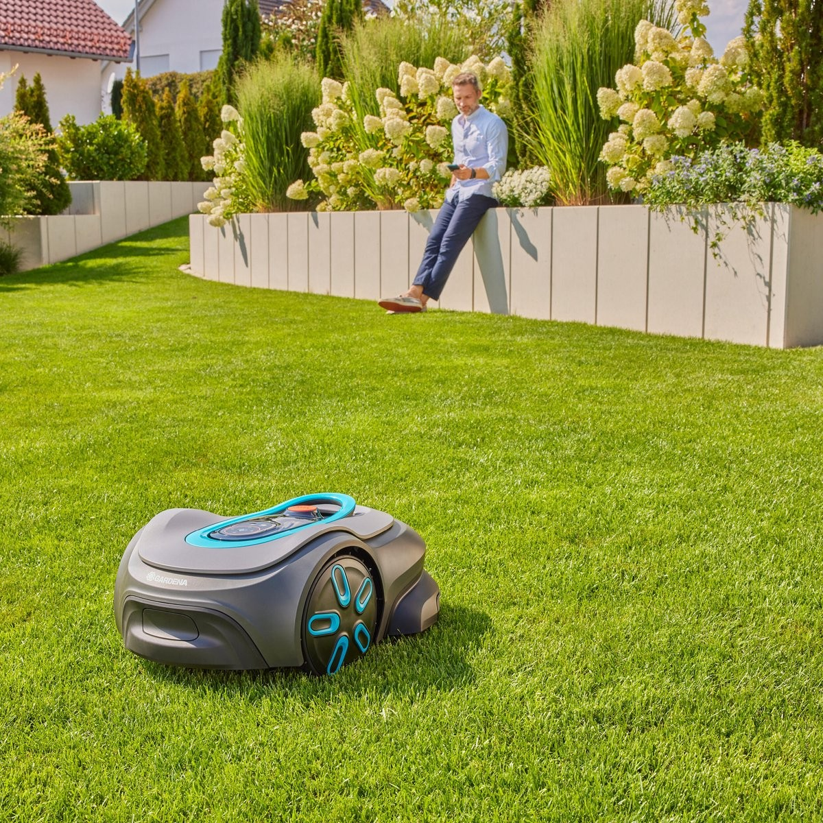 Un hombre sentado en un muro en un jardín junto a un robot cortacésped.