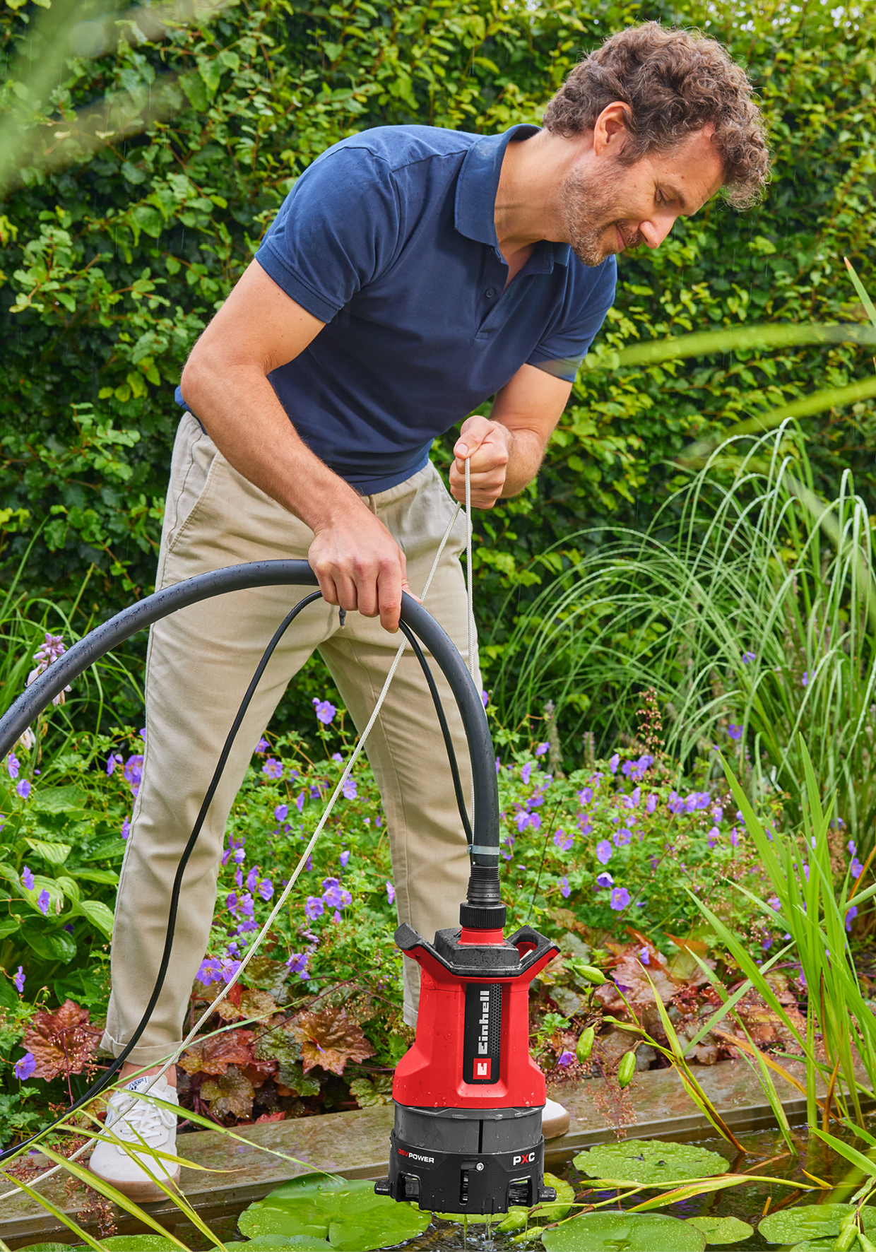 Ein Mann in einem blauen Poloshirt und beiger Hose benutzt eine rote Tauchpumpe im Garten.