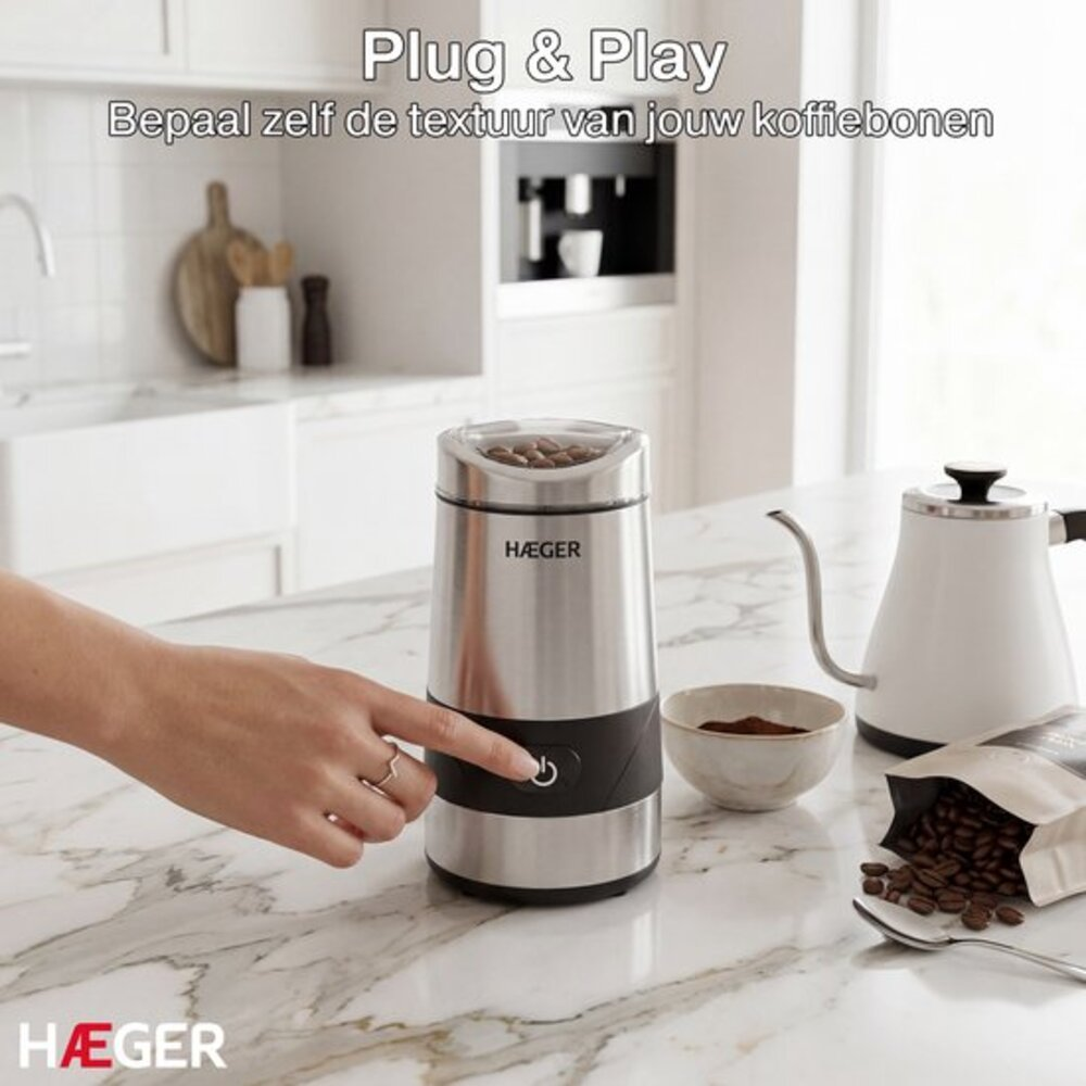 Eine Hand drückt auf einen silbernen Kaffeemühlenknopf. Kaffeebohnen sind sichtbar.