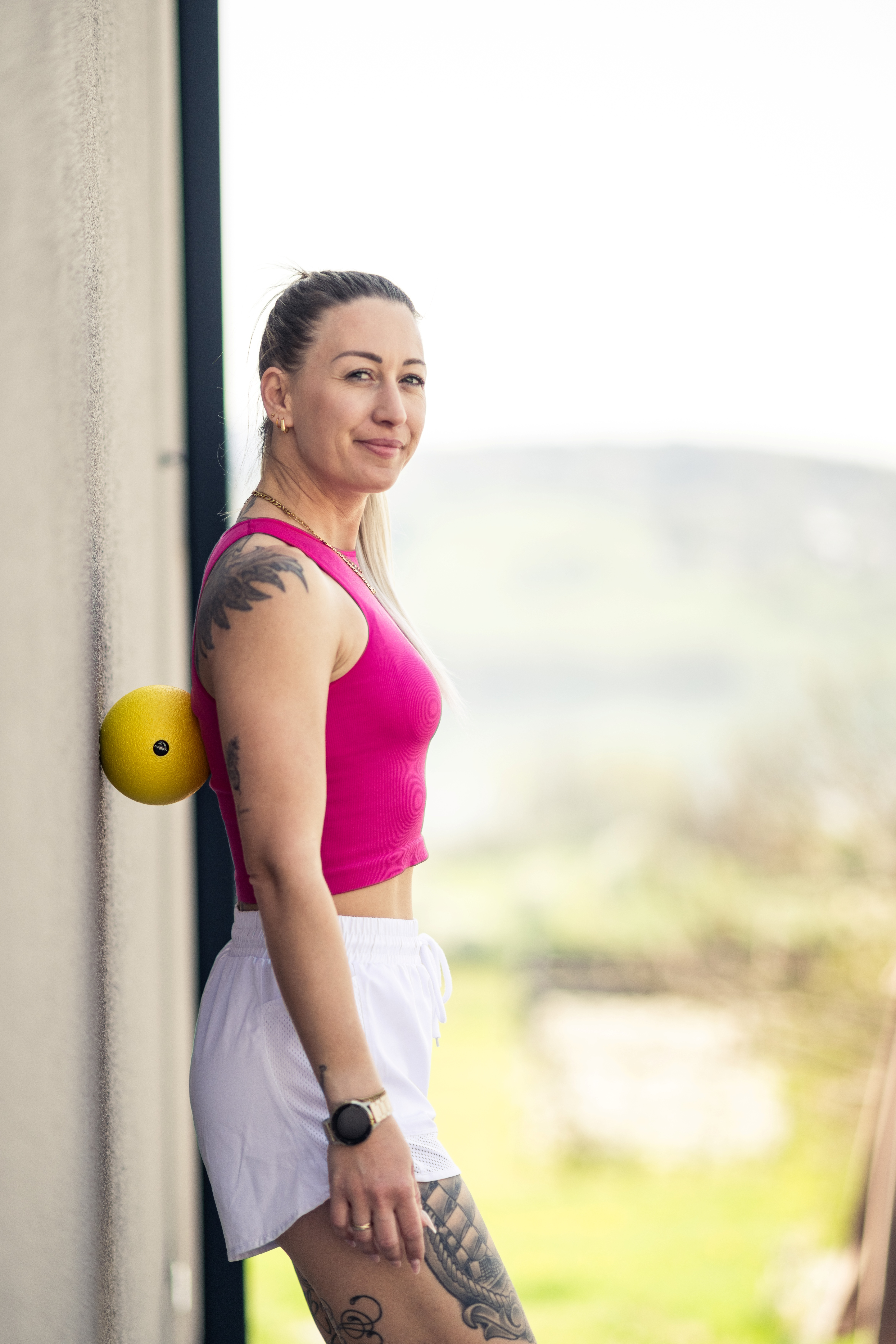 Frau mit Tätowierungen lehnt an einer Wand, ein gelber Ball stützt ihren Rücken.