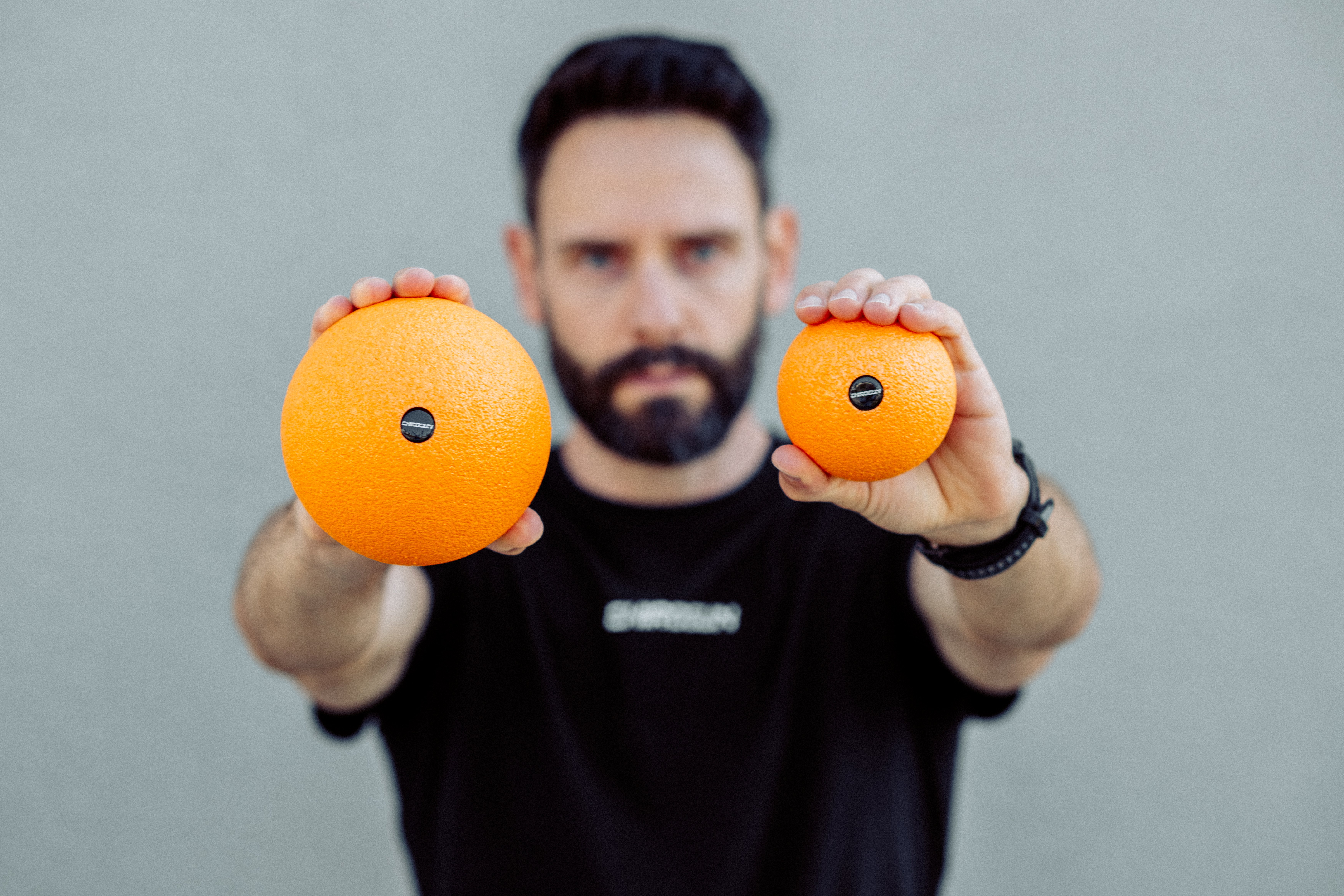 Mann hält zwei orangefarbene Massagebälle vor grauem Hintergrund.