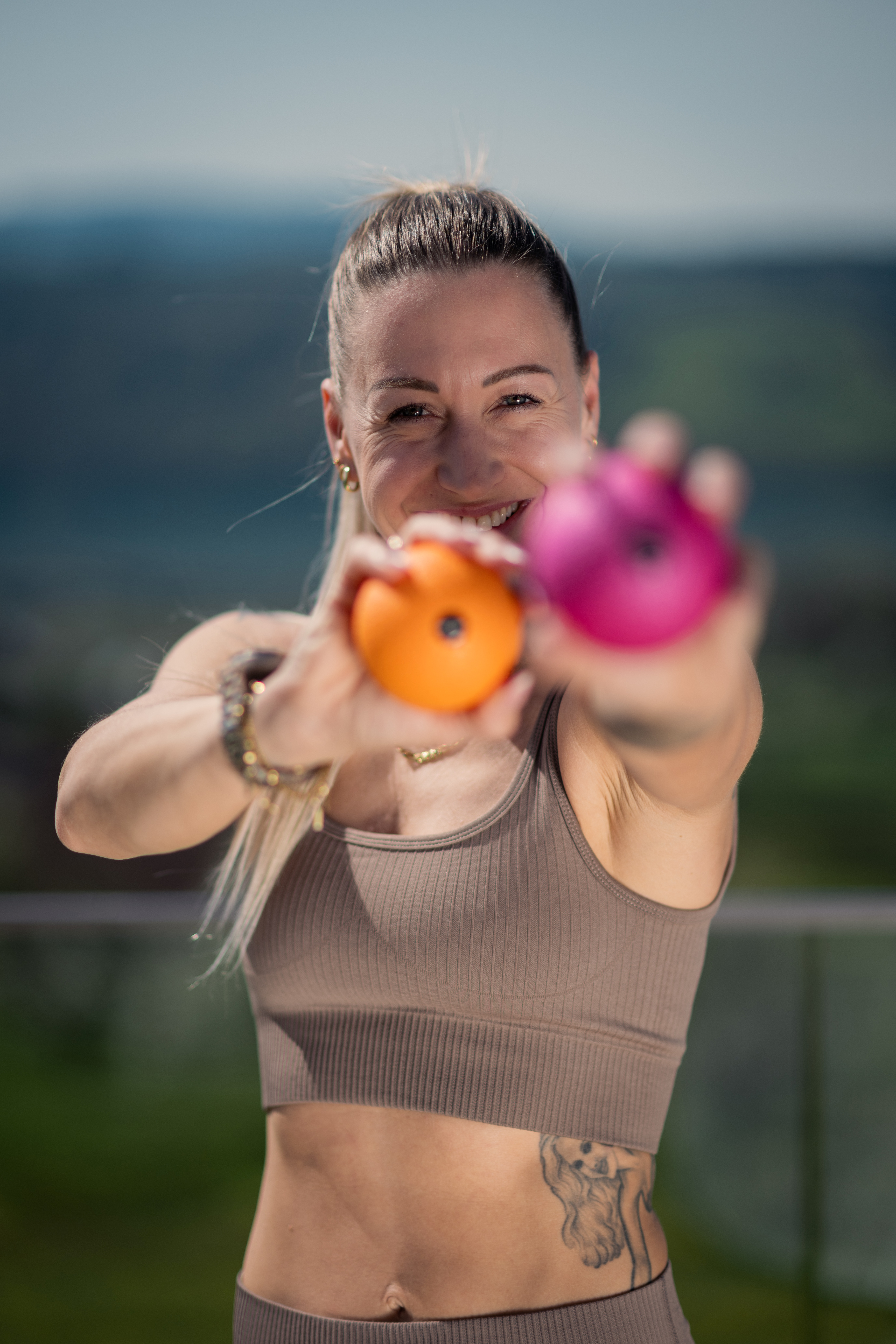 Frau hält orange und pinke Bälle vor sich; trägt Sport-BH; Tattoo am Bauch.