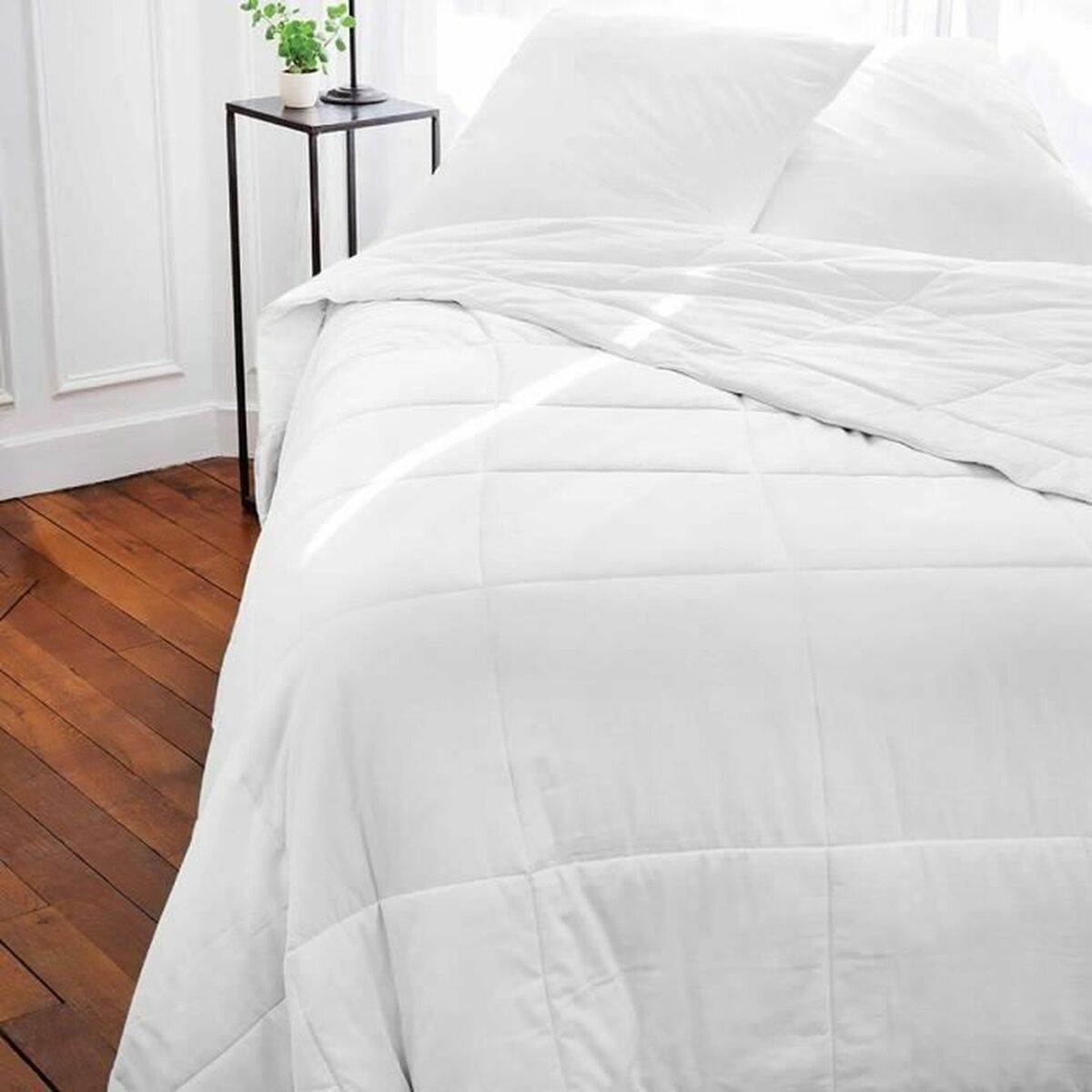 Ein weißes Bett mit Steppdecke, Kissen, Nachttisch und Pflanze.