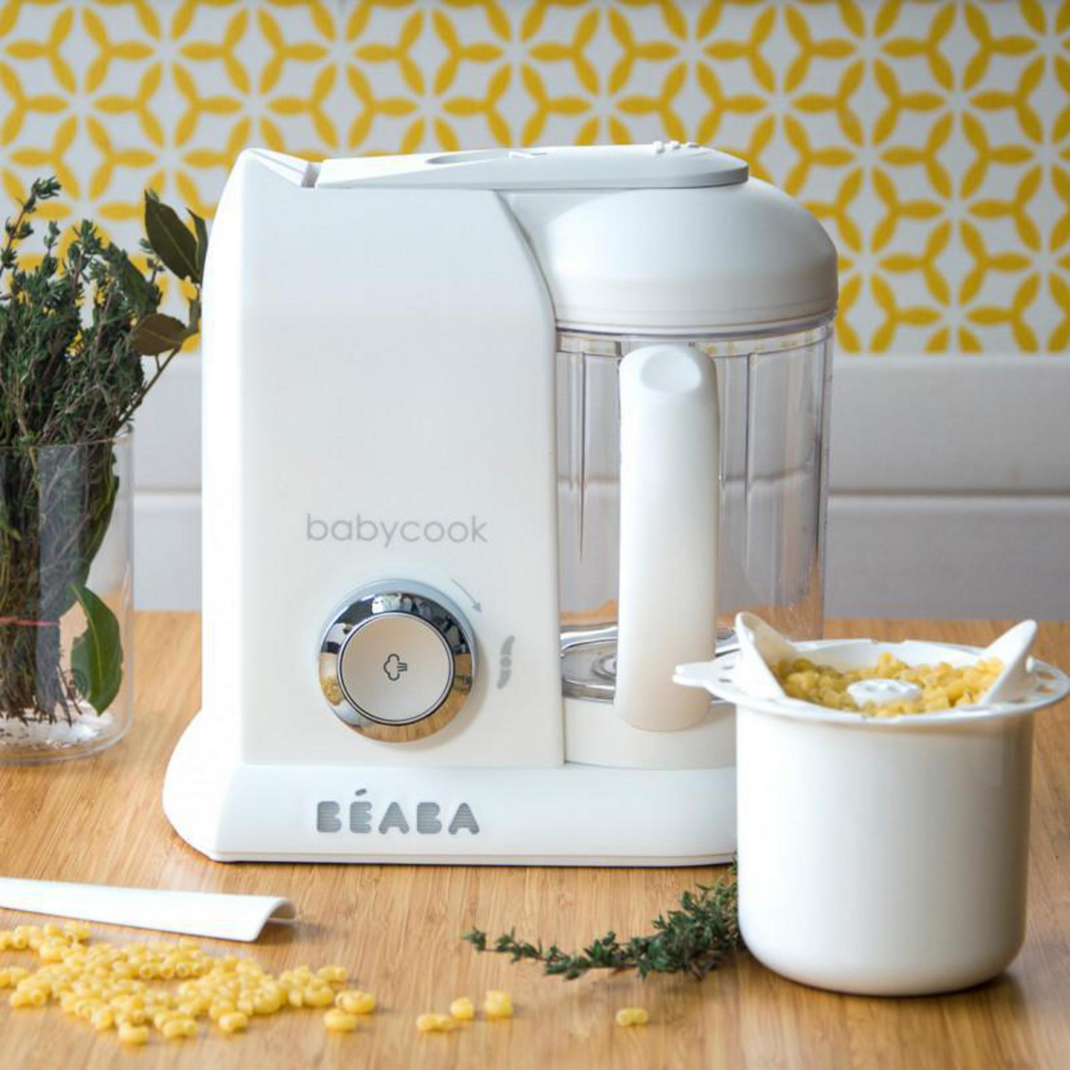 Un vaporizador Babycook blanco sobre mesa de madera con pasta y hierbas.