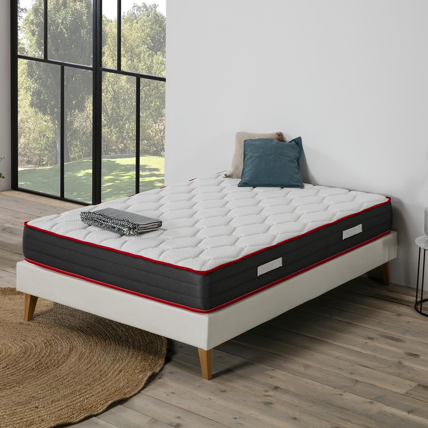 Una cama blanca con colchón, almohadas y manta doblada, cerca de una ventana grande.