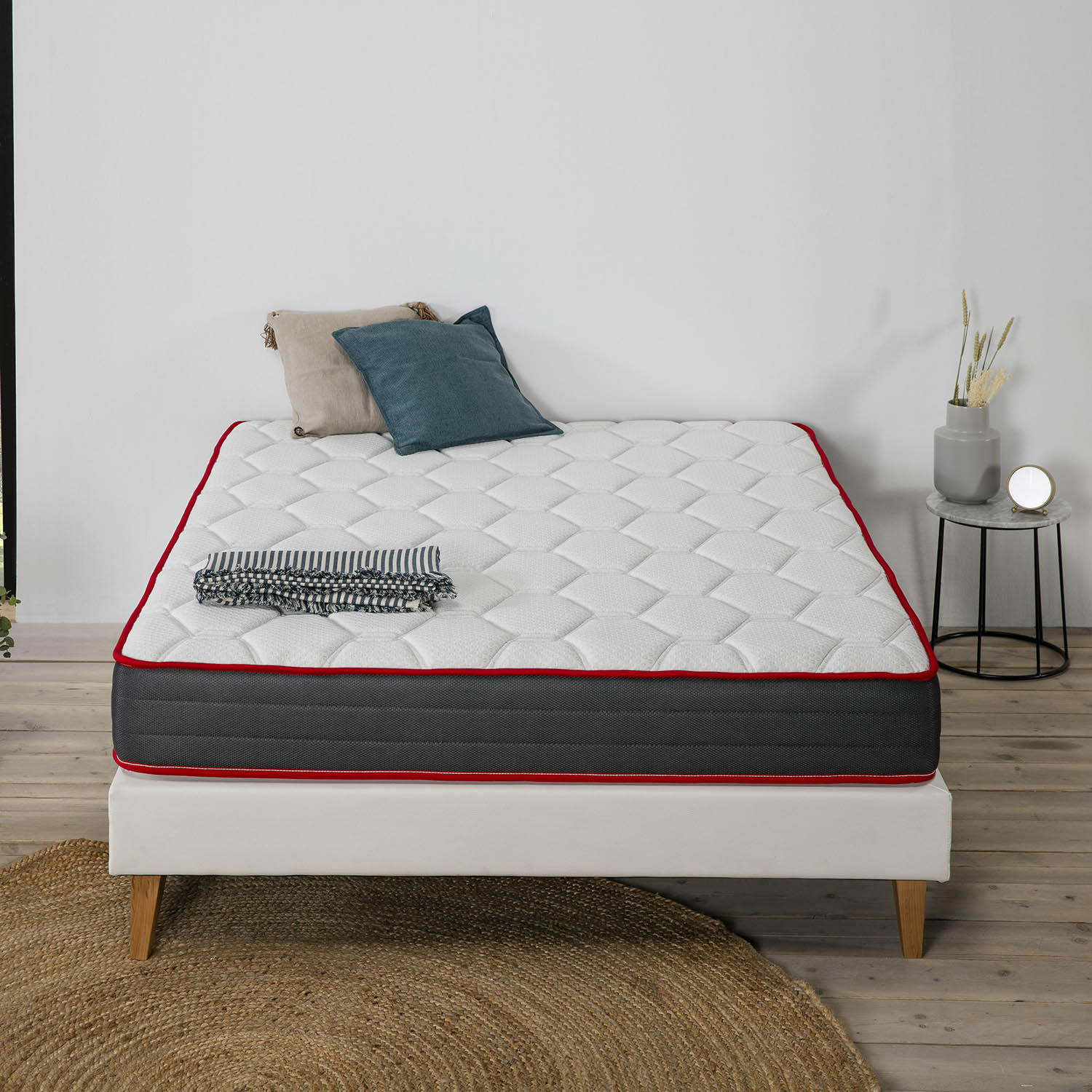 Una cama blanca con colchón, almohadas, manta y mesita auxiliar con jarrón.