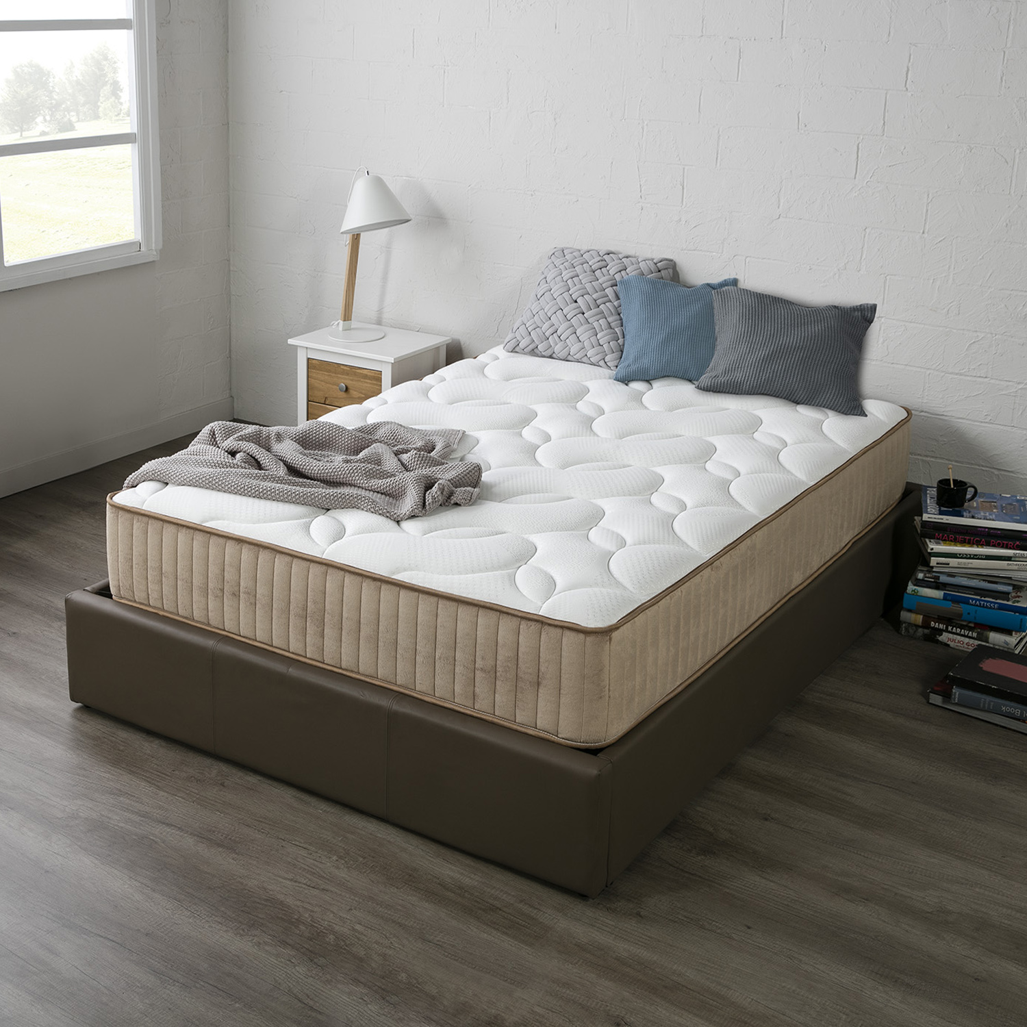 Un colchón con almohadas y manta sobre base de cama, mesita de noche, lámpara y libros.