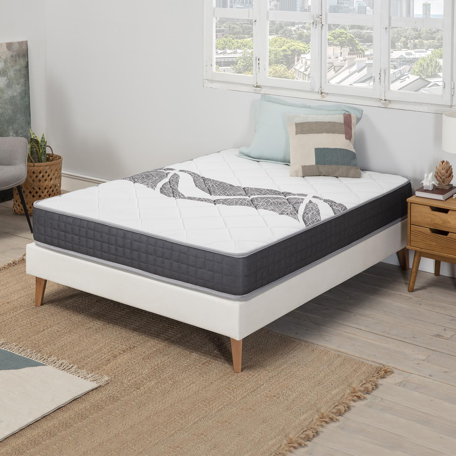 Un colchón blanco con borde gris sobre un somier blanco, con almohadas y mesita de noche.