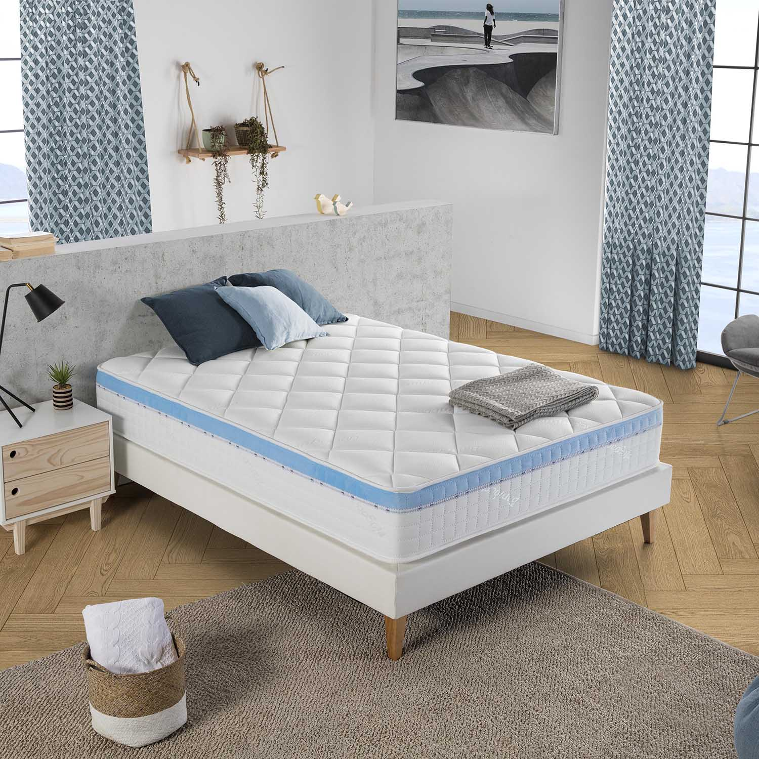 Una cama con colchón blanco, almohadas azules y manta, mesita de noche, cesta con toallas.