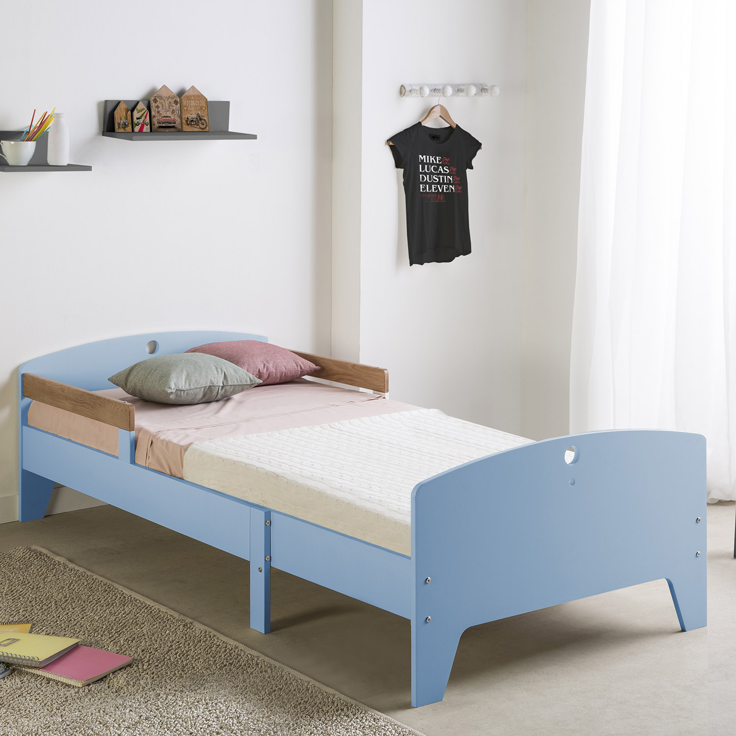 Una cama infantil azul con cojines y manta, al lado una alfombra y libros.