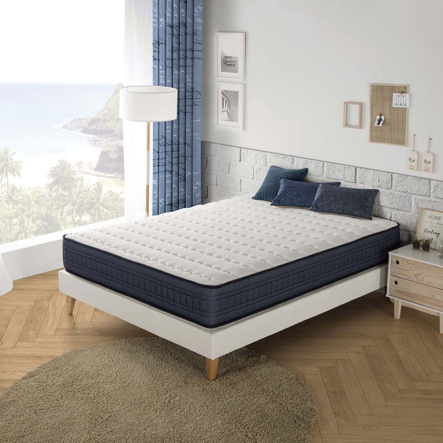 Una cama blanca con colchón, almohadas azules y ventana con vistas al mar.