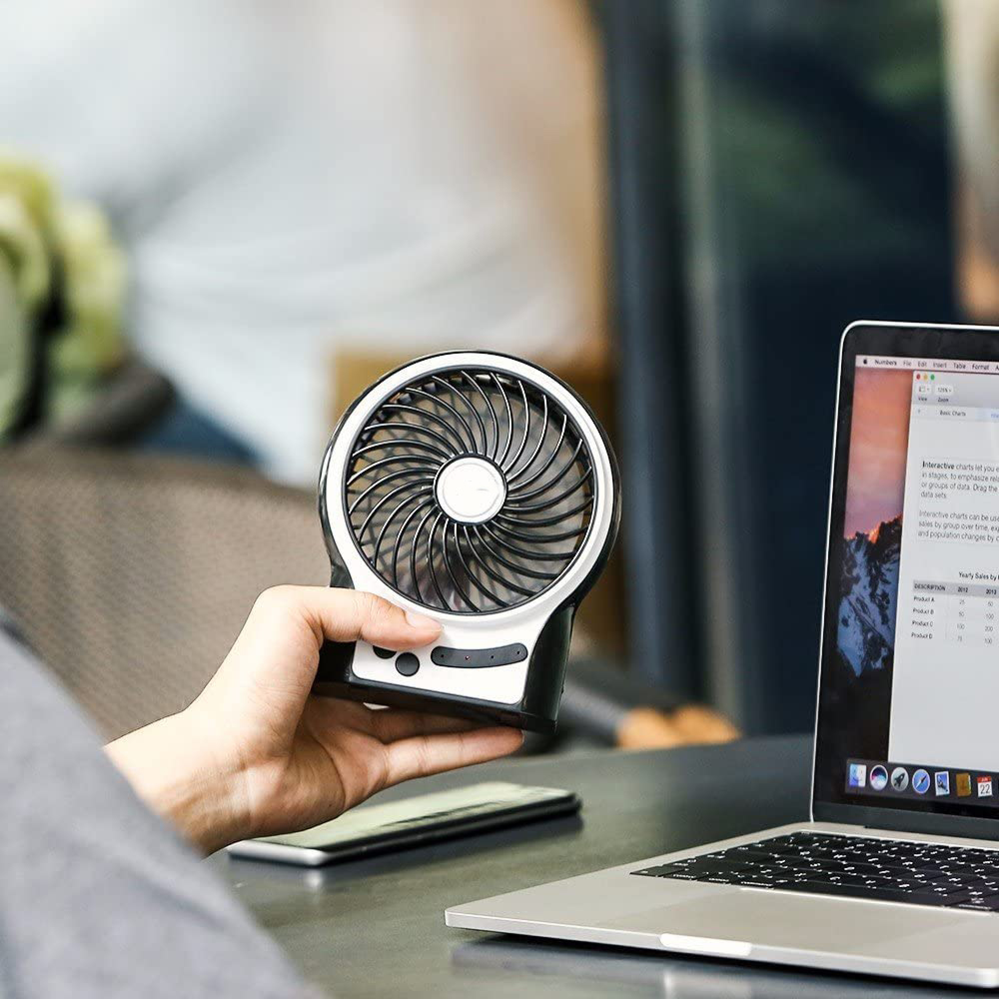 Eine Hand hält einen tragbaren Ventilator neben einem Laptop. Der Ventilator ist schwarz-weiß mit einem runden Gitter.