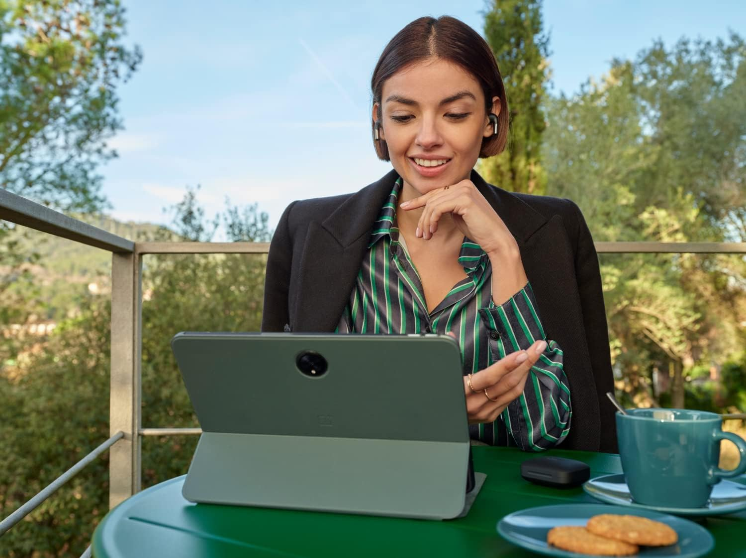 Donna sorridente con auricolari guarda un tablet, all'aperto a un tavolo.