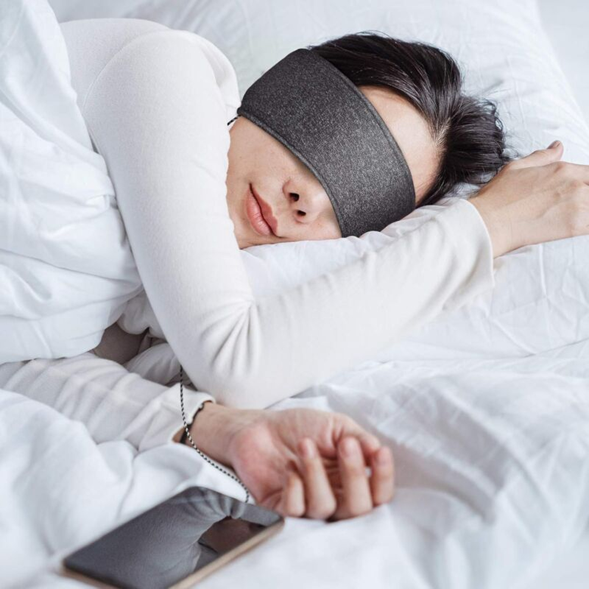 Eine Person schläft auf dem Rücken, trägt eine Schlafmaske und hat ein Handy daneben liegen.