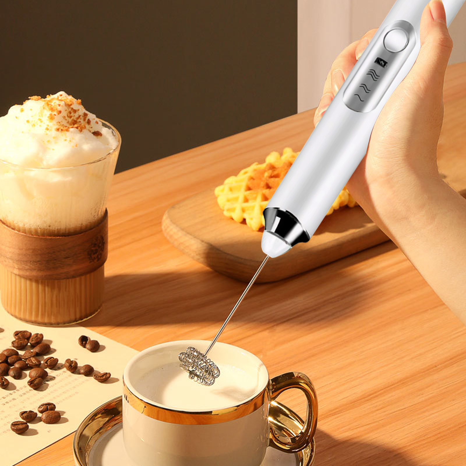 Eine Hand hält einen weißen Milchaufschäumer über einer Tasse, daneben Kaffee und Waffeln.