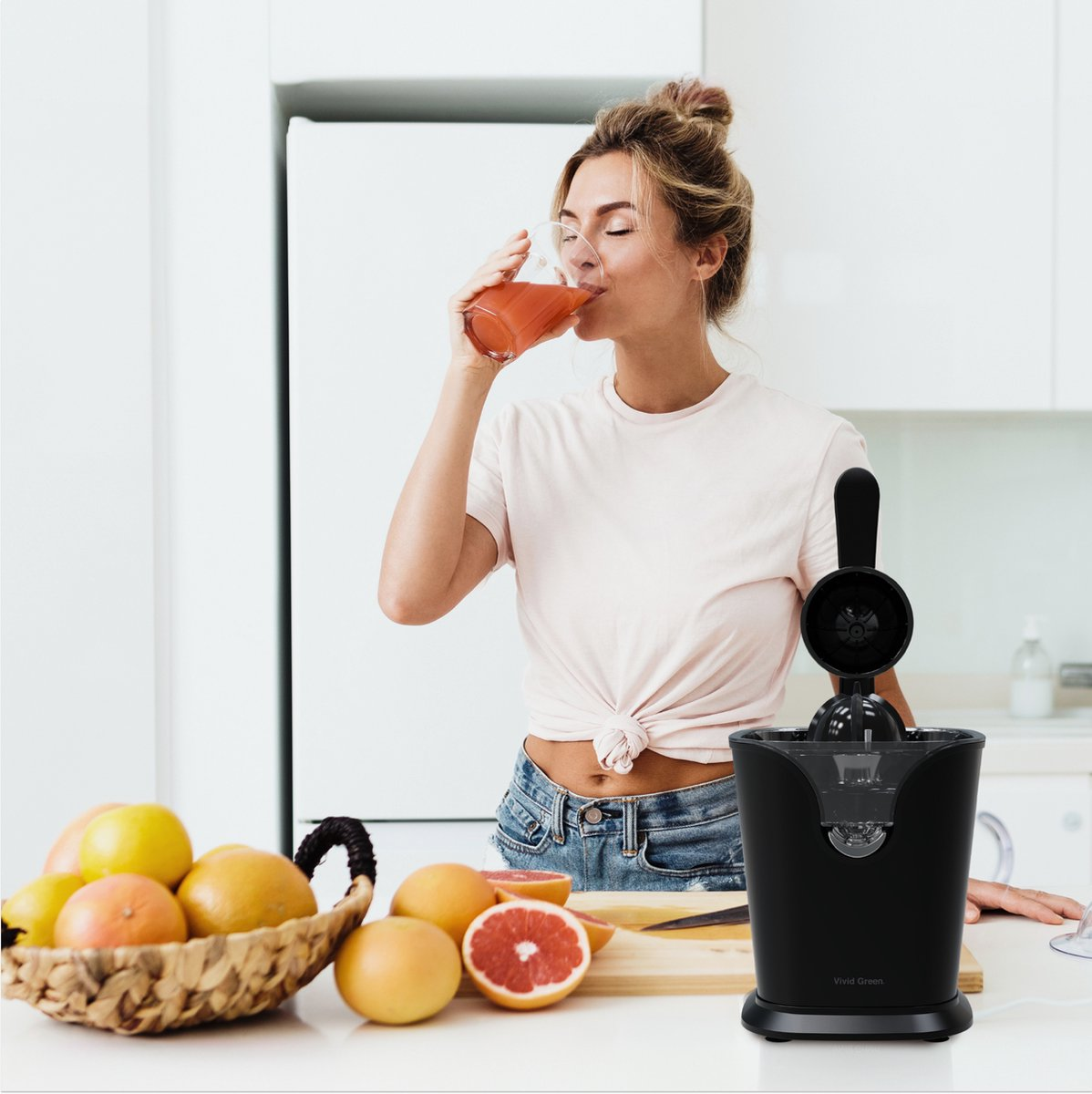 Woman drinks juice in kitchen next to citrus press and fruit.
