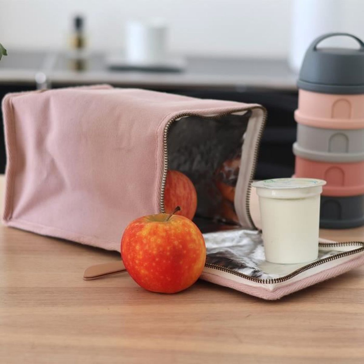 Eine rosa Lunchtasche ist auf einem Holztisch geöffnet, mit zwei Äpfeln und einem Joghurtbecher.