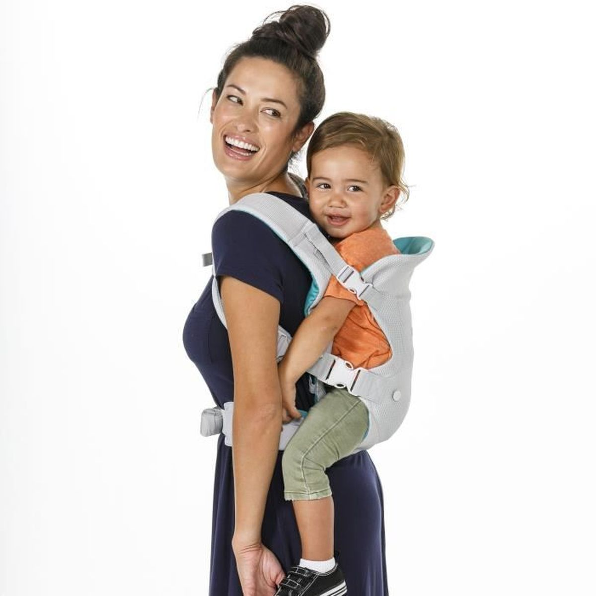 Eine lächelnde Frau in einem marineblauen Kleid trägt ein Baby in einer grauen Trage. Weißer Hintergrund.