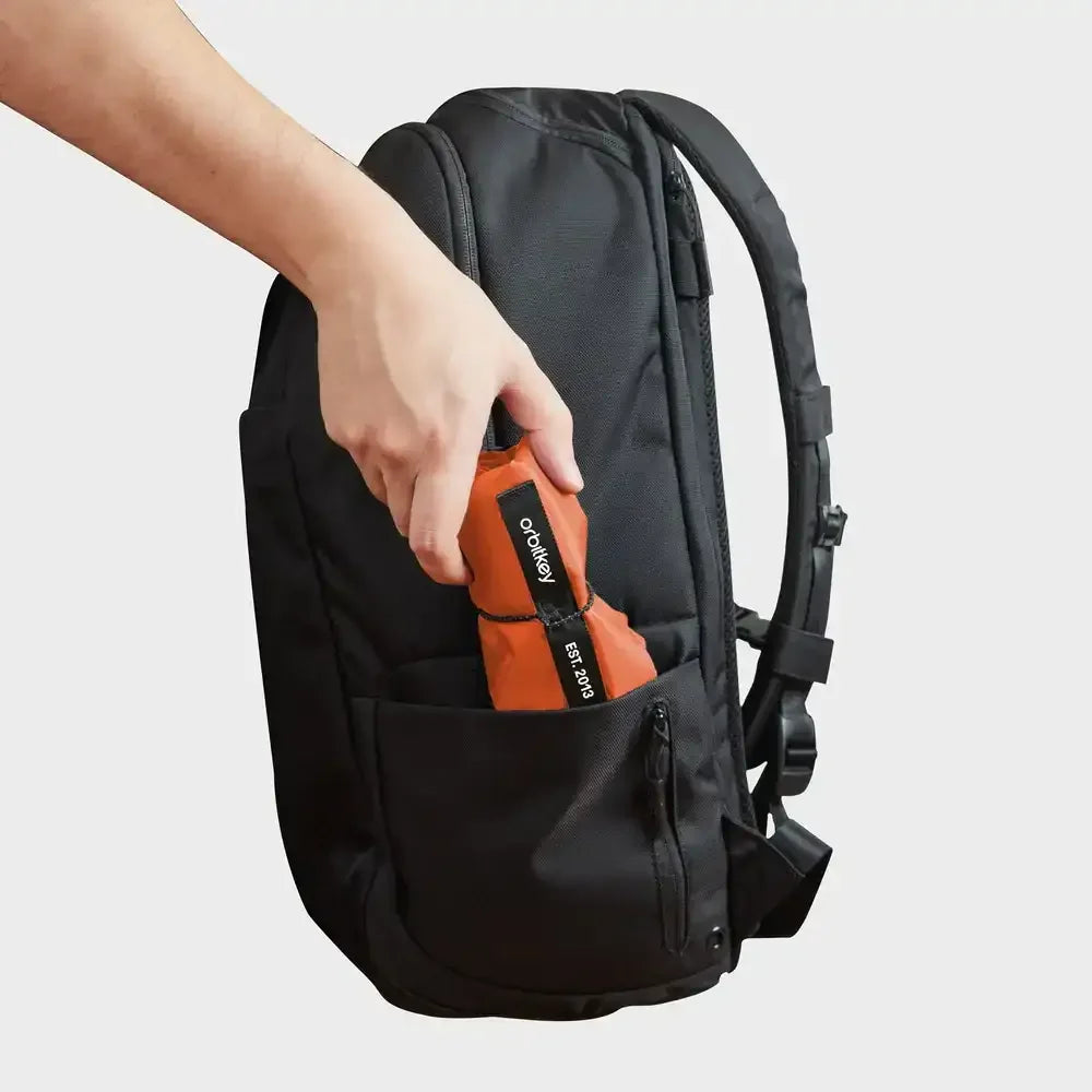 Ręka wkłada pomarańczowy, złożony przedmiot z logo Orbitkey do kieszeni czarnego plecaka.