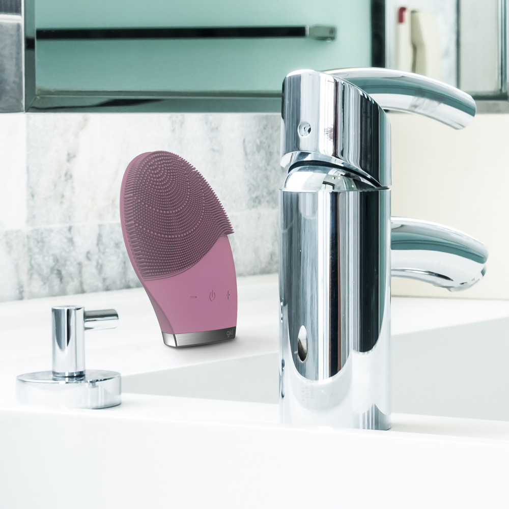 Un cepillo facial rosa yace junto a un grifo brillante en un lavabo.