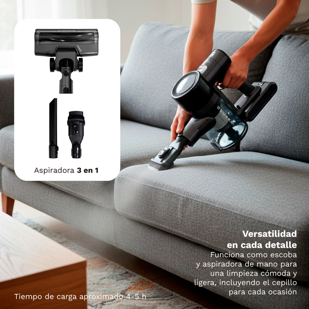 Persona aspira sofá gris con aspiradora negra de mano. Se muestra accesorio.