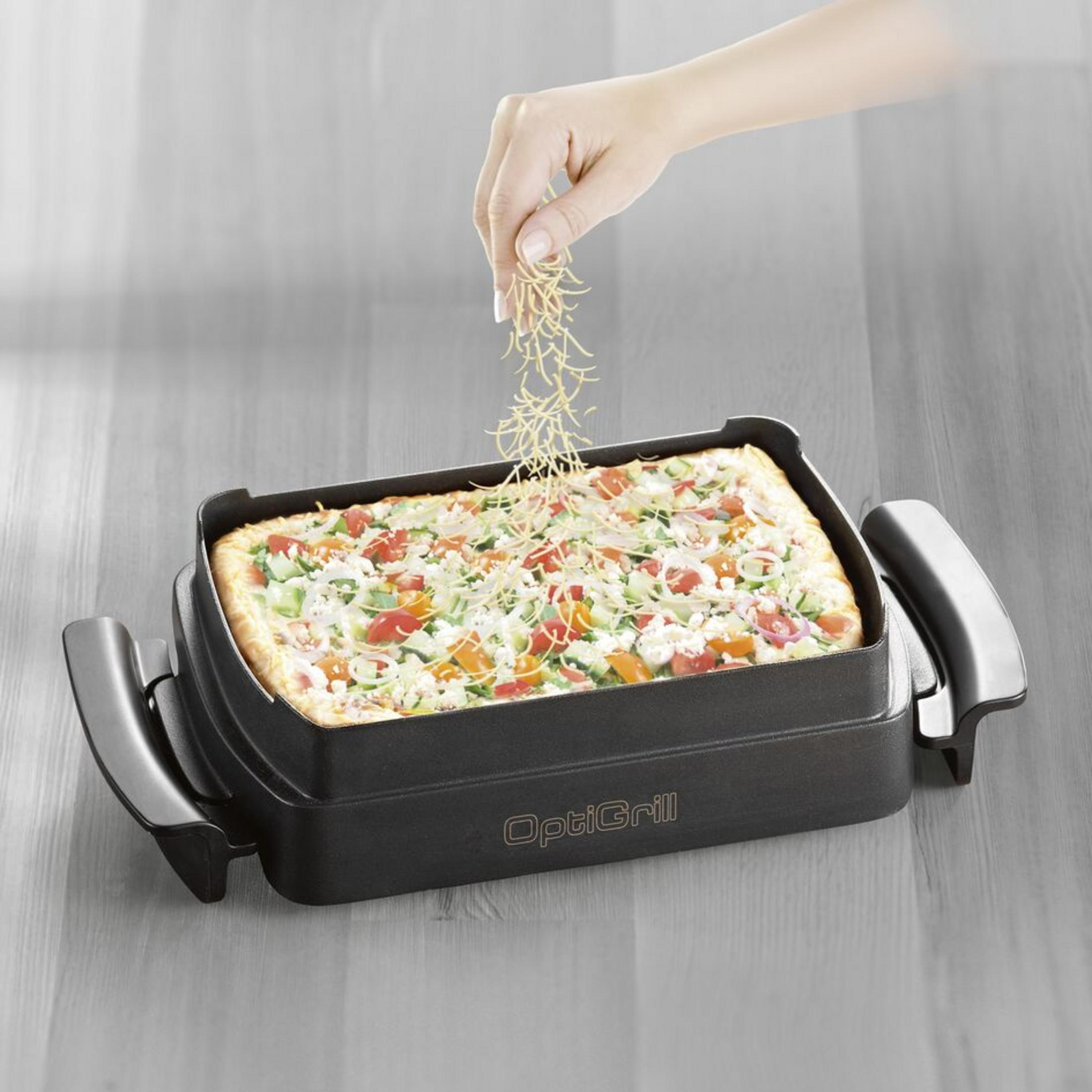 Eine Hand streut Käse über eine rechteckige Pizza in einem schwarzen Ofen.