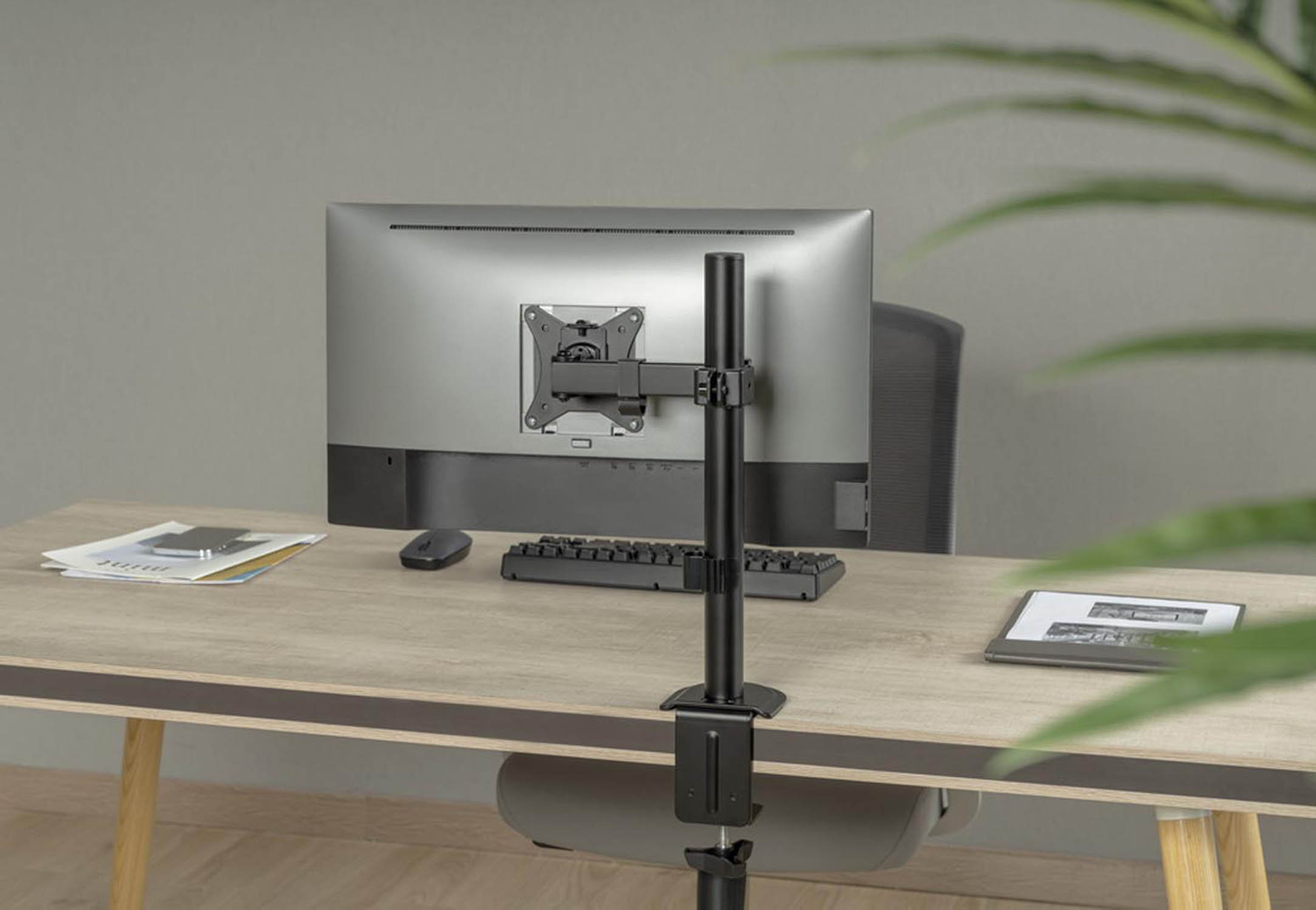 Ein Monitor auf einem Schreibtisch, montiert an einem schwarzen Arm. Tastatur, Maus, Papiere daneben.