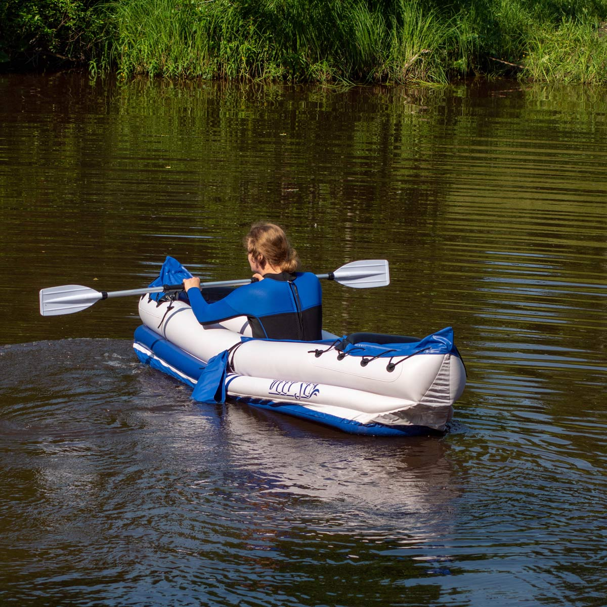 Person paddelt in einem weißen und blauen Kajak auf einem ruhigen Fluss, umgeben von grüner Vegetation.
