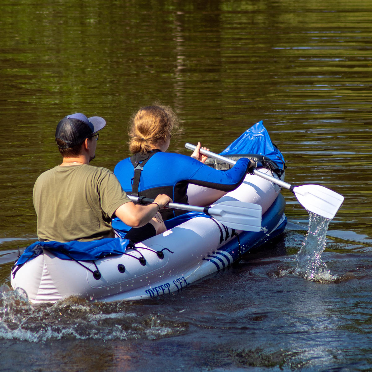 Zwei Personen paddeln mit einem Schlauchkajak auf einem ruhigen See mit grünen Spiegelungen.