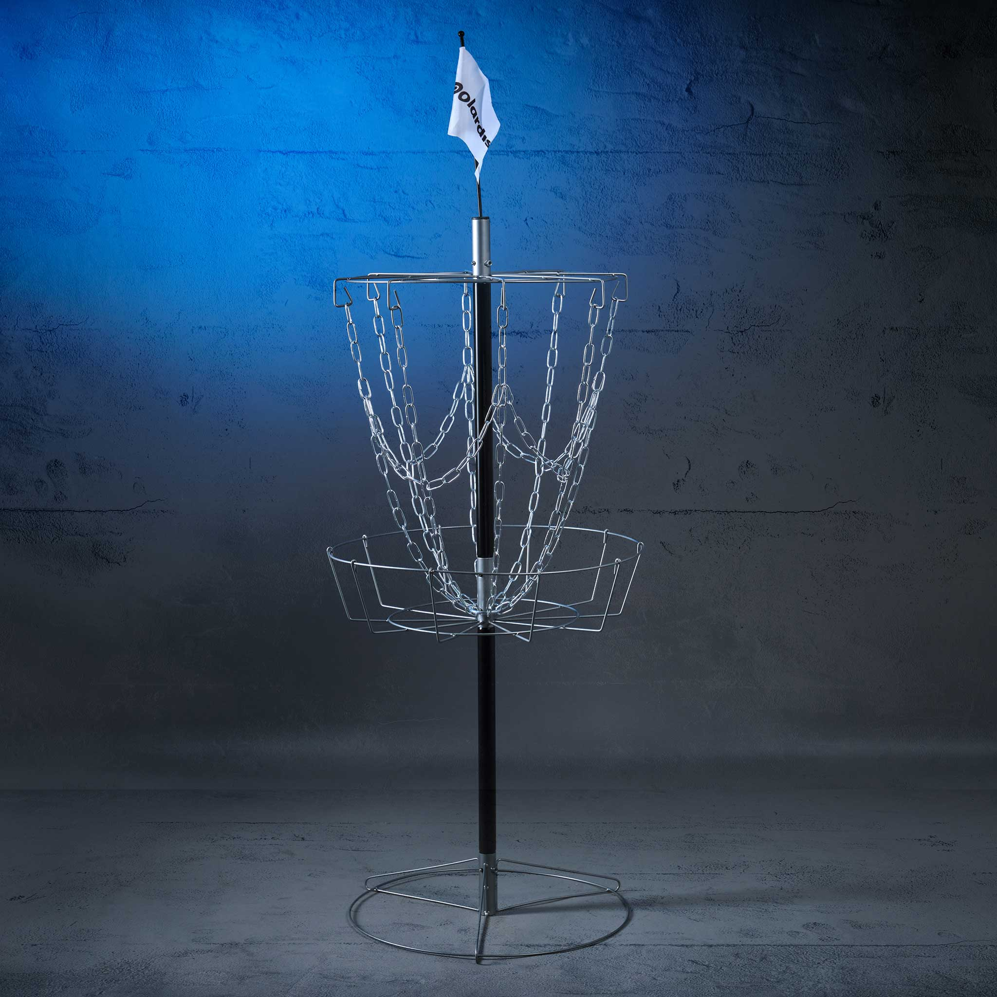 Ein Discgolfkorb mit einer weißen Fahne steht auf einem grauen Boden vor einer blauen Wand.