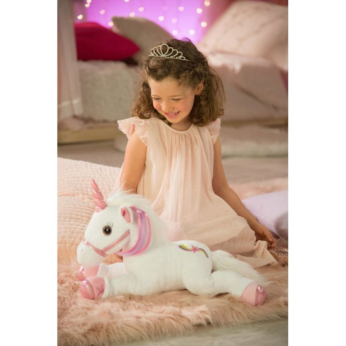 Una niña con tiara y vestido rosa se sienta junto a un unicornio de peluche blanco.