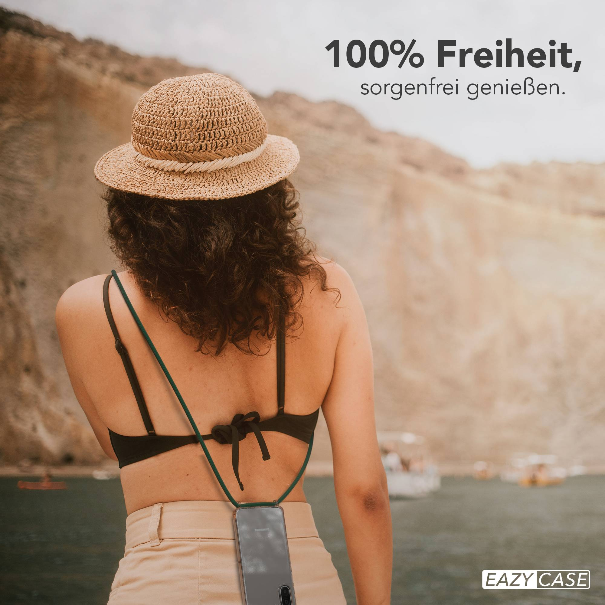 Frau mit Strohhut und Handy-Halterung am Strand, Boote im Wasser.