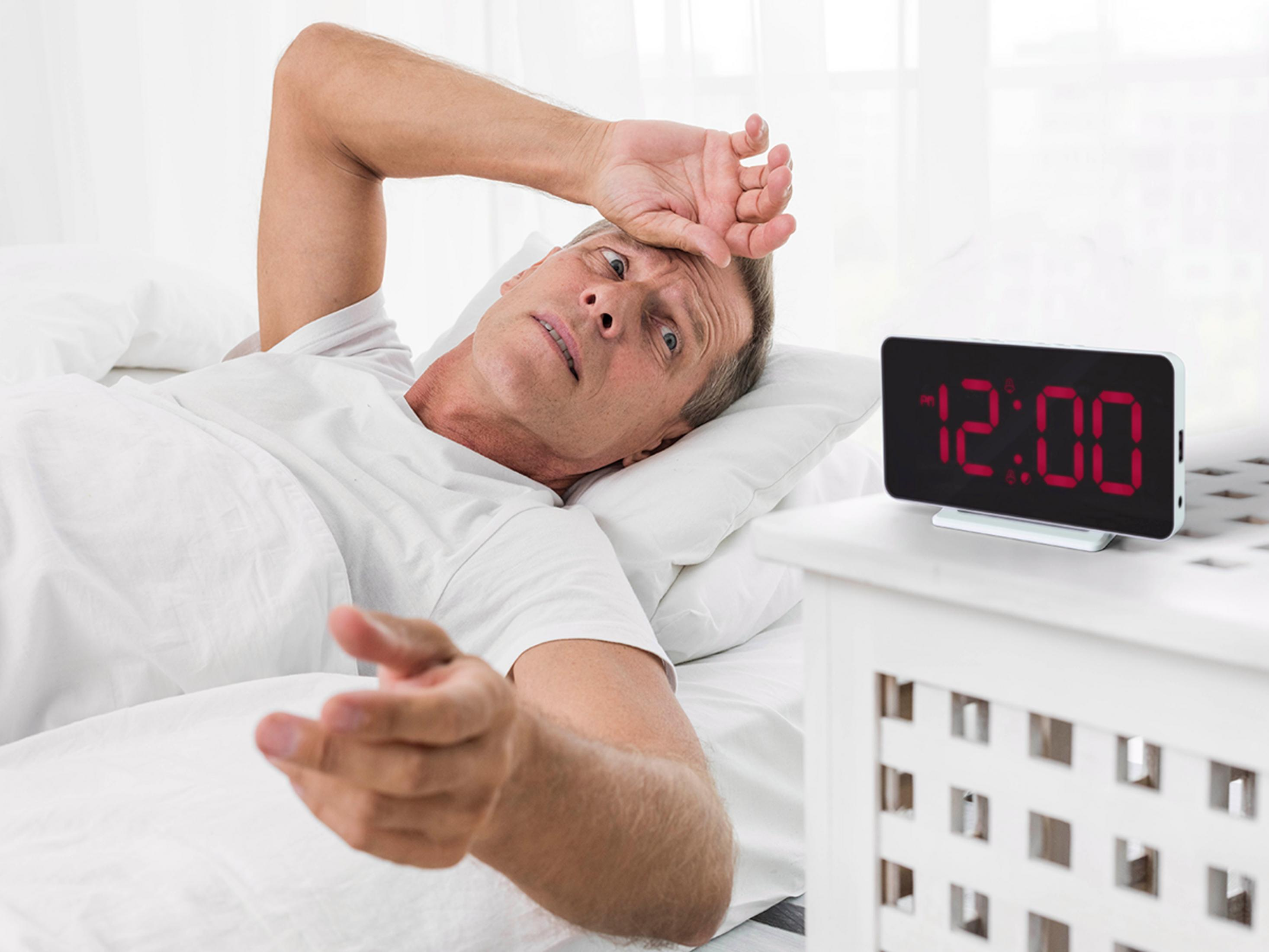 Man lies in bed, holding forehead, digital clock shows 12:00.