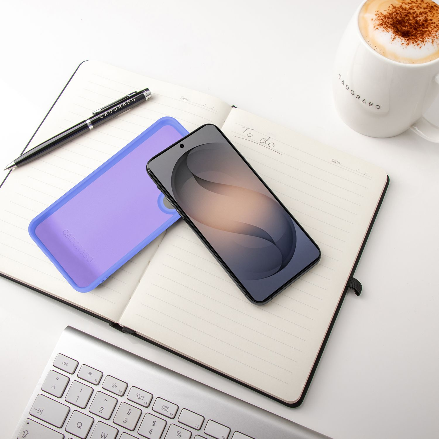 Ein Notizbuch mit Stift, Handy, Tastatur und Kaffee auf einem weißen Schreibtisch.